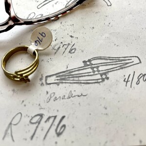 May include: A gold ring with a simple design, a white tag with the number "976" on it, and a drawing of a pair of parallel lines with the number "4/80" written next to it.