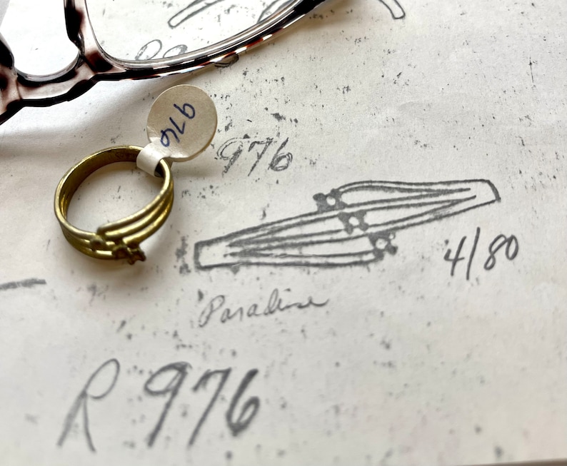 May include: A gold ring with a simple design, a white tag with the number "976" on it, and a drawing of a pair of parallel lines with the number "4/80" written next to it.