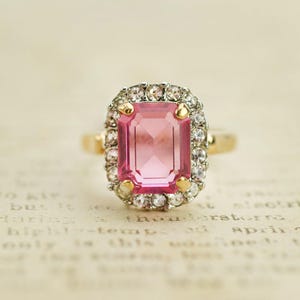 May include: A pink gemstone ring with a square cut stone surrounded by a halo of smaller clear stones.