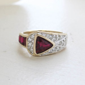 Puede incluir: Un anillo de oro con una gran piedra roja triangular rodeada de piedras claras más pequeñas. El anillo tiene una banda texturizada.