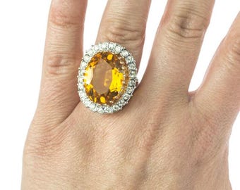 May include: A large, oval-shaped, yellow gemstone ring with a silver setting. The ring is surrounded by smaller, clear gemstones.