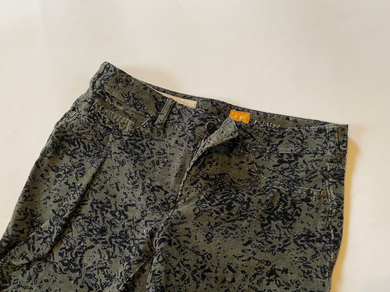 May include: Green corduroy pants with a black abstract print. The pants have a zipper fly and a button closure. The brand name "PILGRIM" is visible on the waistband.