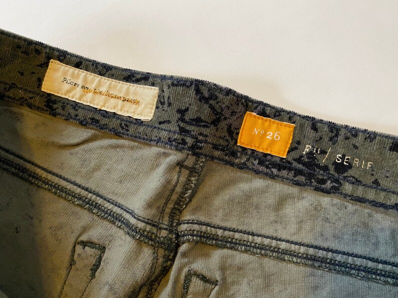 May include: Green corduroy pants with a black and white print. The pants have a label that says "Pilgrim Surf + Supply" and another label that says "No. 26".