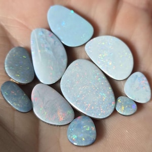 May include: A collection of polished opal gemstones in various shapes and sizes. The opals display a range of colors, including white, blue, and gray, with iridescent flashes. The gemstones are held in a hand, showcasing their natural beauty.