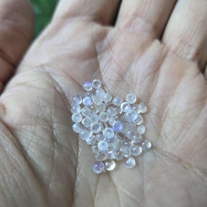 May include: A collection of small, clear, round beads with iridescent, rainbow-like flashes. The beads are held in an open hand, with the light reflecting off their surfaces. The beads appear to be approximately 0.25 inches in diameter, and are scattered across the palm.