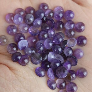 May include: A collection of small, polished amethyst gemstones in varying shades of purple. The gemstones are round and smooth, with a glossy finish. The image shows a close-up view of the gemstones, highlighting their color and texture.