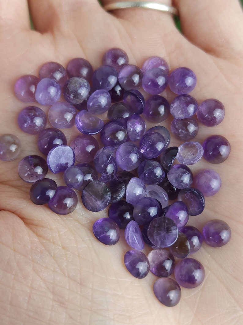 May include: A collection of small, polished amethyst gemstones in varying shades of purple. The gemstones are round and smooth, with a glossy finish. The image shows a close-up view of the gemstones, highlighting their color and texture.