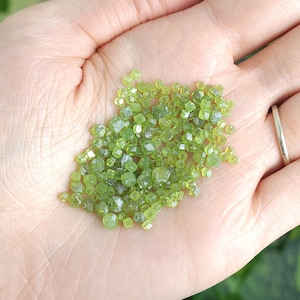May include: A handful of small, light green peridot crystals. The crystals are a vibrant, translucent green and have a faceted, geometric shape. The crystals are held in an open hand, with a silver ring on one finger.