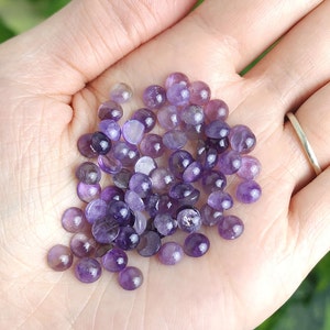 May include: A handful of small, round amethyst gemstones in varying shades of purple. The polished stones are smooth and have a slight sheen, with some showing internal patterns. The gemstones are held in an open hand, with a blurred green background.