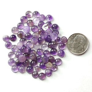 May include: A collection of small, round amethyst gemstones in varying shades of purple, from light lavender to deep violet. A U.S. dime is included for size reference. The gemstones are smooth and polished, with a cabochon cut.