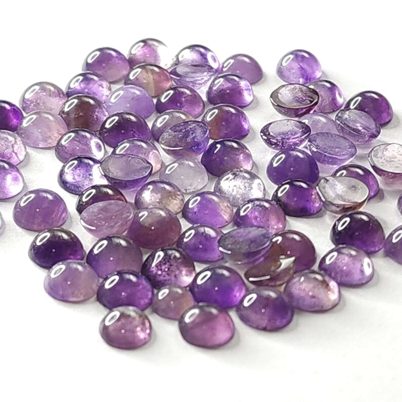 May include: A collection of polished, cabochon-cut amethyst gemstones in varying shades of purple. The gemstones are oval-shaped and have a smooth, rounded surface. The background is white, highlighting the color and clarity of the stones.