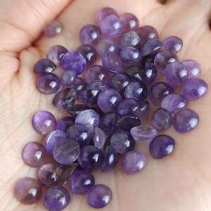 May include: A collection of polished, round amethyst gemstones in varying shades of purple. The gemstones are smooth and have a slightly domed shape, with a glossy finish. The stones are held in a hand, showcasing their natural color and texture.