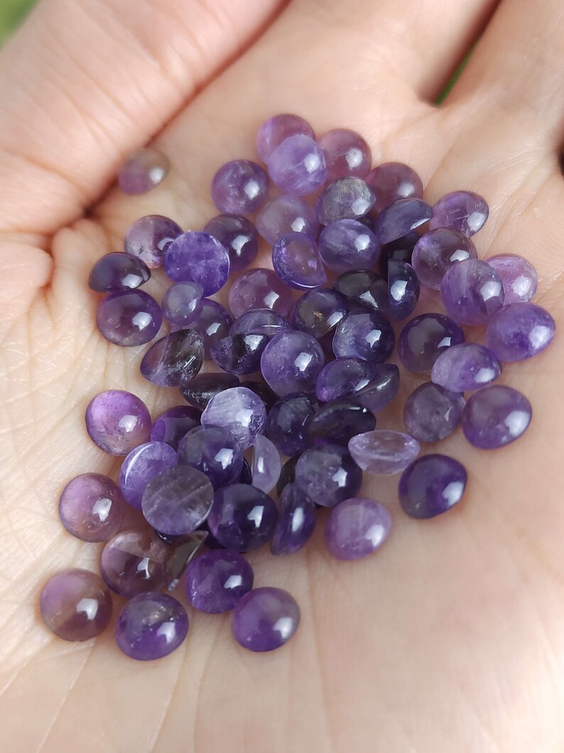 May include: A collection of polished, round amethyst gemstones in varying shades of purple. The gemstones are smooth and have a slightly domed shape, with a glossy finish. The stones are held in a hand, showcasing their natural color and texture.