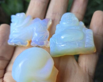 May include: Three opalescent crystal carvings: a skull, a seated Buddha, and a figure. The carvings are held in a hand, showcasing their translucent, iridescent quality. The Buddha carving is seated, and the other figure is standing.