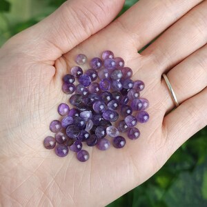 May include: A handful of small, polished amethyst gemstones in varying shades of purple. The gemstones are round and smooth, with a slight dome shape. The gemstones are held in an open palm, with a silver ring visible on one finger.