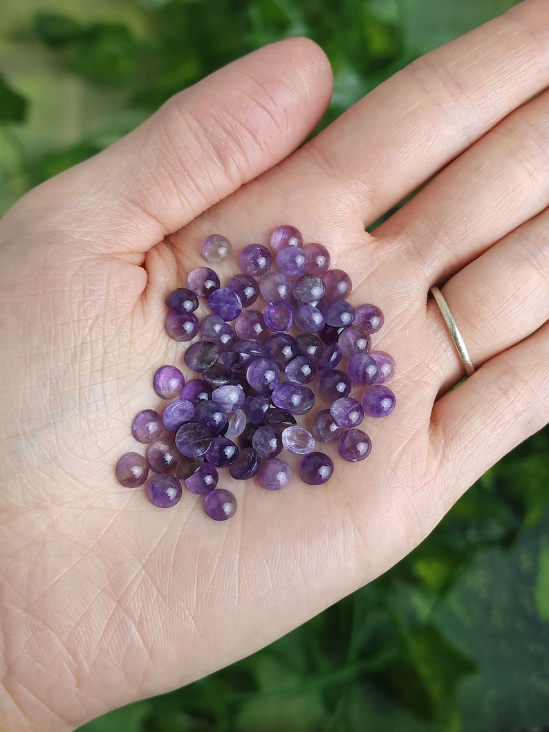 May include: A handful of small, polished amethyst gemstones in varying shades of purple. The gemstones are round and smooth, with a slight dome shape. The gemstones are held in an open palm, with a silver ring visible on one finger.