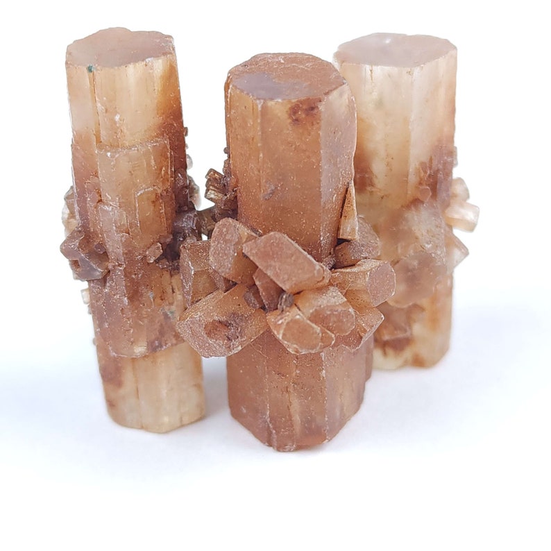 May include: Three aragonite crystal clusters. The crystals are a warm, earthy brown color with a translucent quality. The crystals have a columnar structure with clusters of smaller crystals around the base and sides. The background is white.
