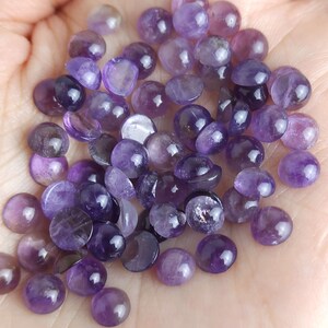 May include: A collection of polished amethyst cabochons in various shades of purple, from light lavender to deep violet. The gemstones are round and smooth, with a slight dome shape, and are displayed in a hand.