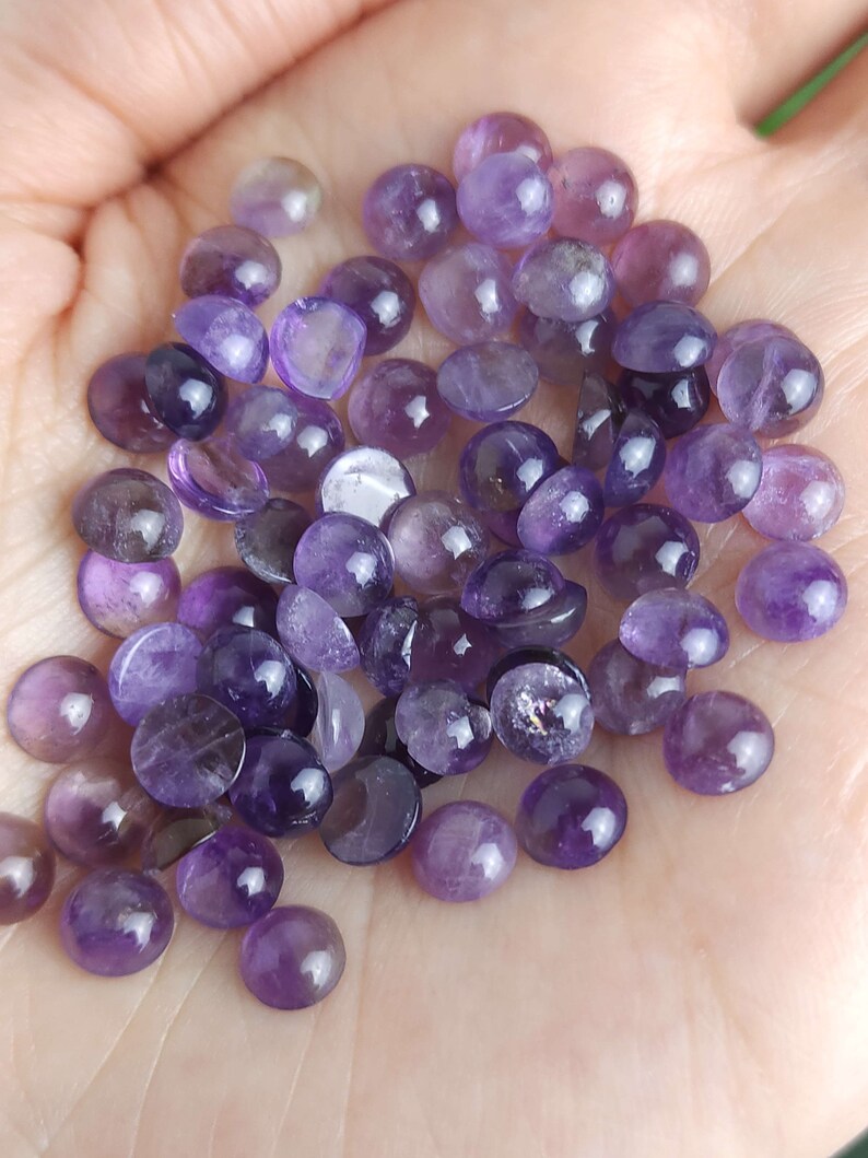 May include: A collection of polished amethyst cabochons in various shades of purple, from light lavender to deep violet. The gemstones are round and smooth, with a slight dome shape, and are displayed in a hand.