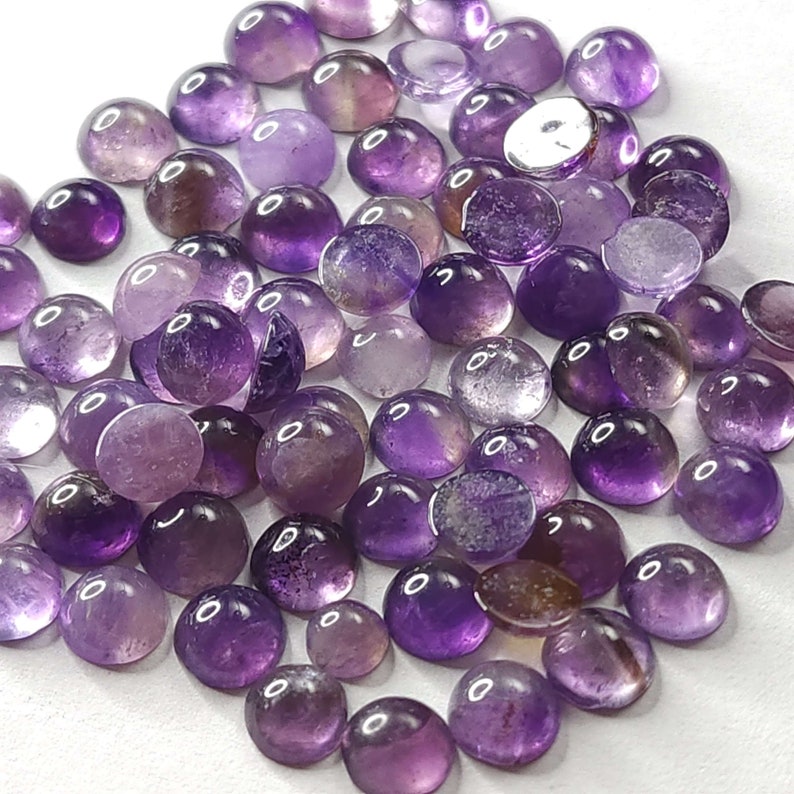 May include: A collection of polished amethyst cabochons in various shades of purple, from light lavender to deep violet. The gemstones are round and domed, with a smooth, glossy surface. The image shows a close-up view of the gemstones, highlighting their color variations and clarity.