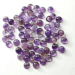 May include: A collection of round, polished amethyst gemstones in varying shades of purple. The gemstones are scattered across a white surface, showcasing their translucent quality and natural color variations. These cabochons are ideal for jewelry making.