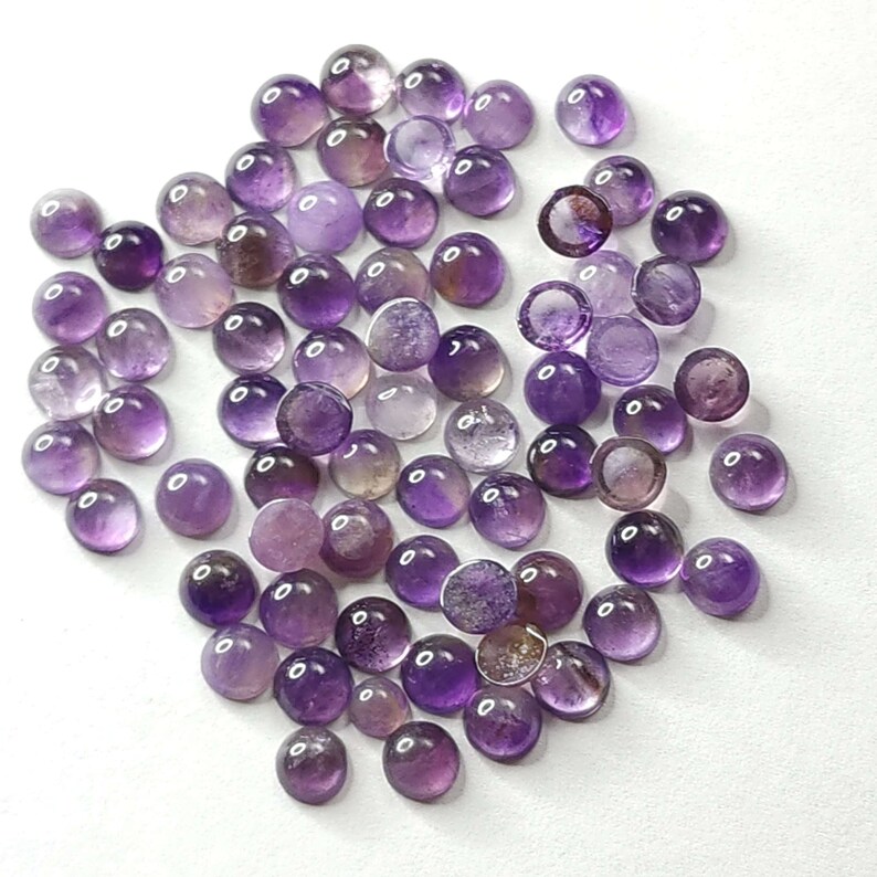 May include: A collection of round, polished amethyst gemstones in varying shades of purple. The gemstones are scattered across a white surface, showcasing their translucent quality and natural color variations. These cabochons are ideal for jewelry making.