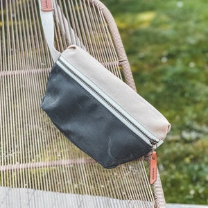 Natural and Charcoal Grey  color block waxed canvas fanny pack with russet leather accents, draped over the back of a chair.