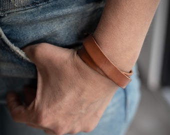 May include: A light brown leather bracelet wrapped twice around the wrist. The bracelet is a simple, minimalist design. The person is wearing blue jeans and a gray t-shirt.