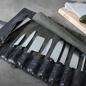 以下が含まれることがあります： キャンバスフラップが付いた黒い革製のナイフロールで、8本のナイフが入っています。ロールが開いており、ナイフとそのハンドルが見えています。ナイフは一列に並んでおり、刃は上を向いています。