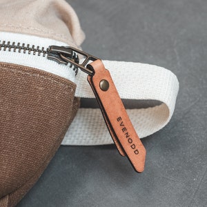 Close up of a Natural and Tan color block waxed canvas fanny pack, featuring an antique brass zipper with russet leather zipper pull secured with an antique brass rivet