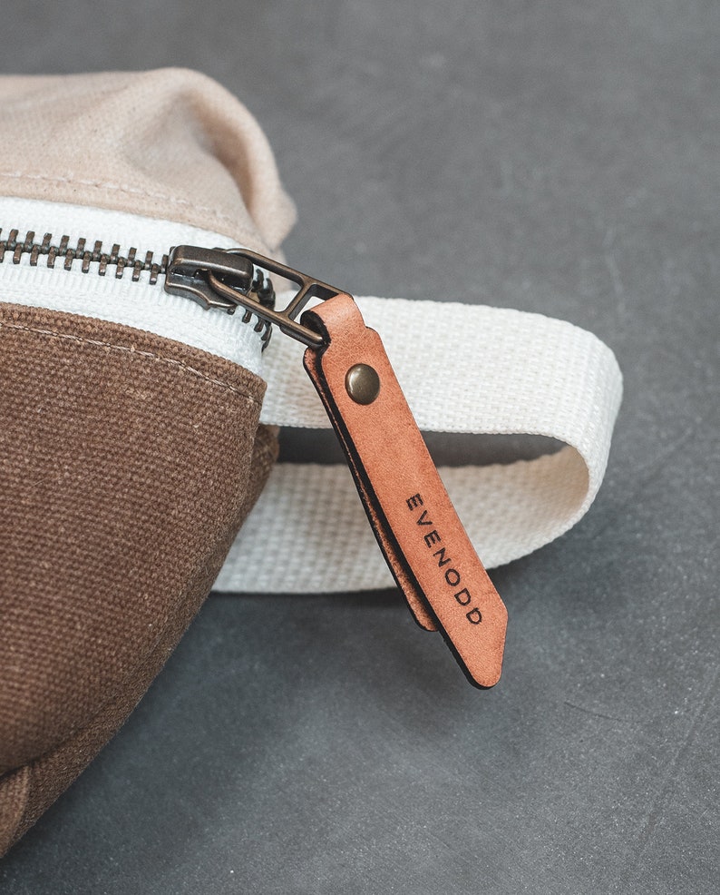 Close up of a Natural and Tan color block waxed canvas fanny pack, featuring an antique brass zipper with russet leather zipper pull secured with an antique brass rivet