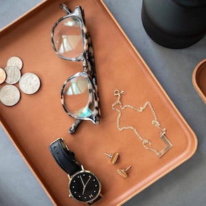 Puede incluir: Una bandeja de cuero marrón con un par de gafas de carey, un collar de cadena dorada con un colgante de barra, un par de pendientes de oro y un reloj de pulsera negro con una esfera dorada.