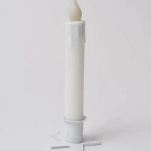 Puede incluir: Una vela LED blanca con una llama parpadeante realista, colocada en una base blanca en forma de estrella. La vela tiene un efecto de cera goteada. La vela mide aproximadamente 25 cm de altura.