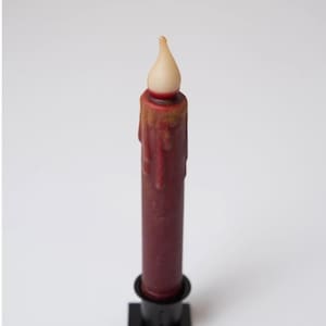 Könnte beinhalten: Eine weinrote elektrische Kerze mit einem realistischen Wachstropfendesign. Die Kerze hat eine cremefarbene Flamme und steht in einem schwarzen quadratischen Sockel. Die Kerze ist so gestaltet, dass sie wie eine traditionelle Kerze aussieht.