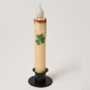 May include: A cream-coloured battery-operated candle with a faux flame, featuring a green shamrock design. It sits in a black metal candlestick holder. The top of the candle has a brown, wax-like appearance, reminiscent of a real candle.