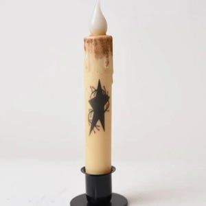 May include: A cream-coloured battery-operated candle featuring a black star design. The top of the candle has a brown, textured appearance, and it sits in a black metal holder. The candle's flame is a soft white colour.