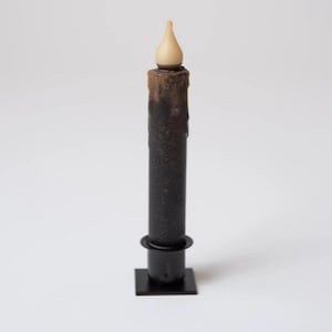 May include: A black, battery-operated taper candle with a faux flame. The candle has a textured finish with brown wax drips. It stands on a black base with a square bottom.