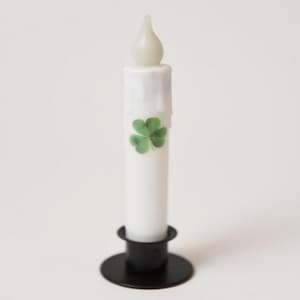 May include: A white LED candle with a green shamrock design, set in a black metal candle holder. The candle has a flame-shaped tip and a dripped wax effect.