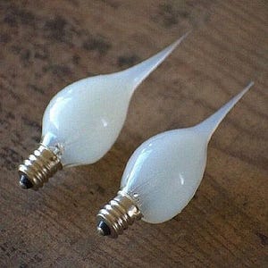 May include: Two white, flame-shaped light bulbs with brass-colored bases and black tips. The bulbs are on a wooden surface. The bulbs have a candelabra base.