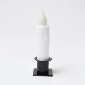 May include: A white LED candle with a flickering flame effect, set in a black metal candlestick holder. The candle has a realistic wax appearance with subtle drips. The base of the holder is square.