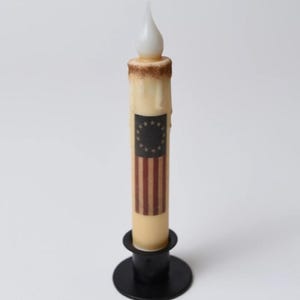 Puede incluir: Una vela que funciona con pilas con un cuerpo de cera de color crema y una llama LED parpadeante. La vela presenta un diseño impreso de la bandera estadounidense y está colocada en un candelabro de metal negro. La vela mide aproximadamente 20 cm de altura.