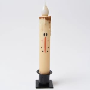May include: A snowman-themed LED candle with a cream-colored body and a brown top. The candle features a painted face with black eyes, a red nose, and black dots. It sits in a black metal holder.