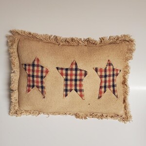 May include: Rectangular burlap pillow with three plaid stars in red, navy, and white. The pillow has a fringed edge. The pillow is a decorative accent for a home.