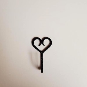 May include: A black metal wall hook featuring a decorative top shaped like a heart with two curved scrolls meeting at the center. The hook has a smooth, dark finish and a simple, functional design that extends downward from the heart-shaped base.