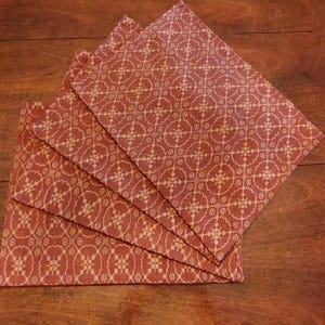 May include: Four folded red and cream-colored cloth napkins with a repeating geometric pattern. The napkins are arranged in a fan shape on a wooden surface, showcasing their design and texture.