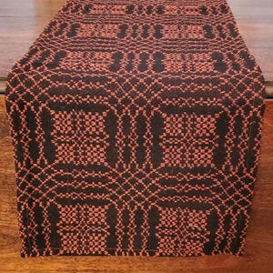 May include: A rectangular table runner featuring a woven geometric pattern in contrasting black and burnt orange threads. The textile displays a repeating grid-like design with intricate interlocking shapes across the entire surface area.