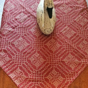 Pode incluir: Um caminho de mesa tecido em vermelho e branco com um padrão geométrico repetitivo de diamantes. Uma figura decorativa de cisne branco com bico preto e marcas pretas desgastadas está centrada no tecido, colocado sobre uma superfície de madeira.