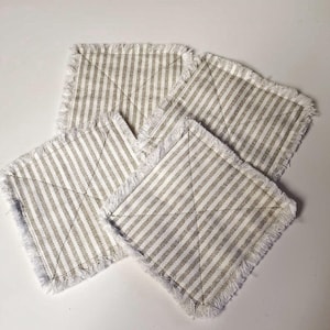 May include: Four square coasters with a striped pattern in beige and white. Each coaster has a fringed edge. The coasters are arranged in a slightly overlapping manner, showcasing their design and texture. These coasters are suitable for home decor.