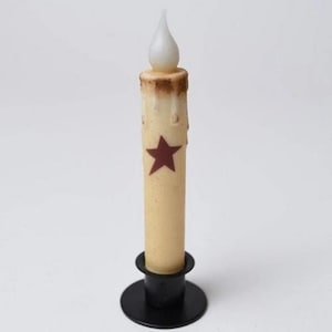 May include: A battery-operated candle with a cream-colored body and a red star. The candle has a realistic flickering flame and sits in a black metal base. The candle has a rustic, aged appearance.