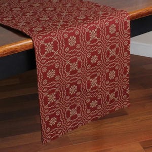 May include: A red and tan patterned table runner draped over a wooden table. The runner features a repeating geometric design in tan against a red background. The runner's edge hangs over the table's edge.